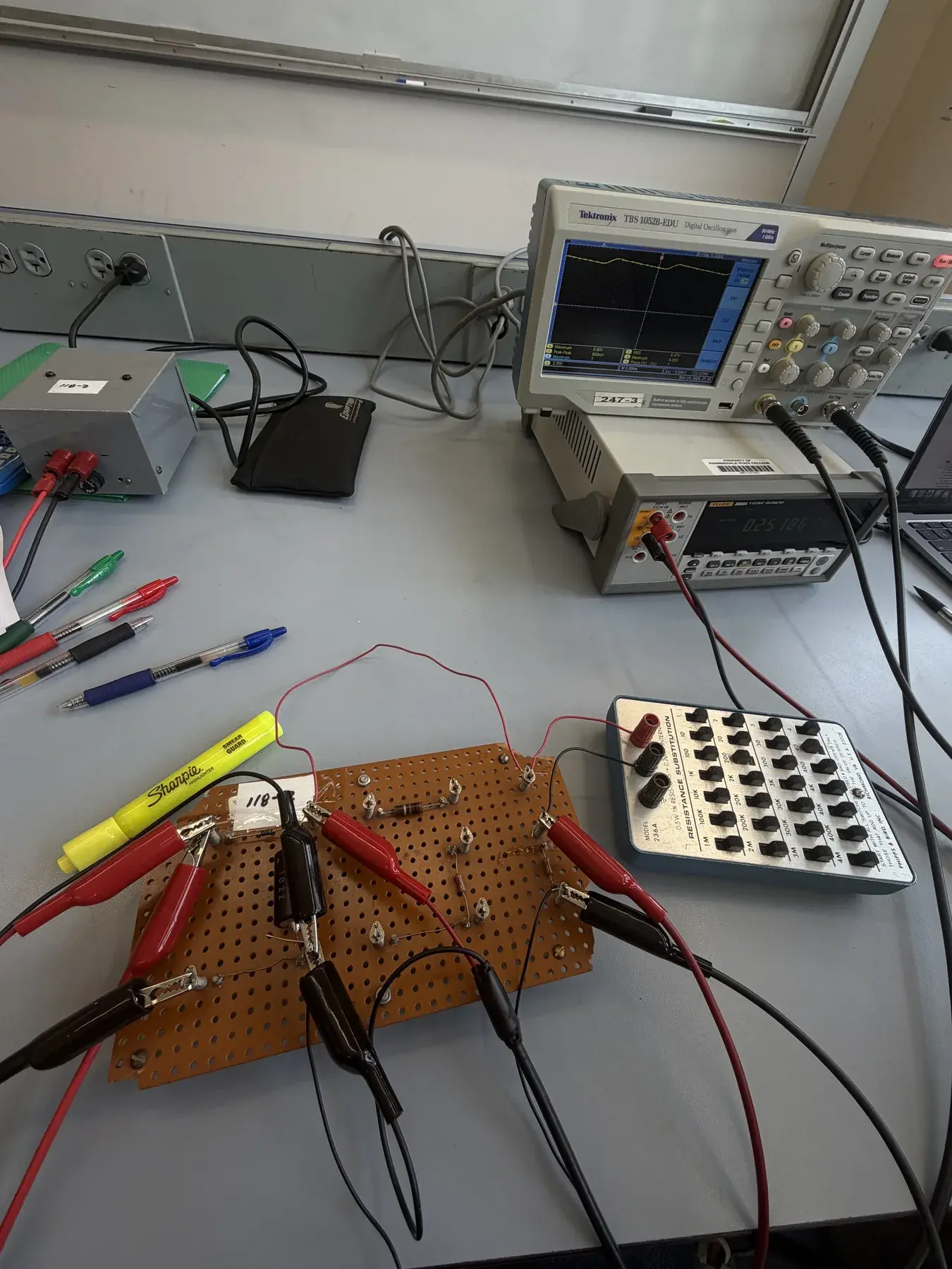 Electronics bench with a perforated prototyping board, diodes, resistor, capacitor, test leads, oscilloscope, multimeter, and resistance substitution box